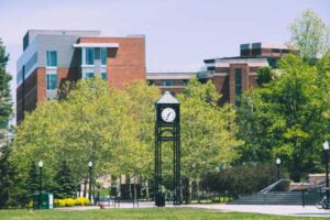 A large clock tower stands tall on a sidewalk near a grassy field on the University of Rochester's campus.